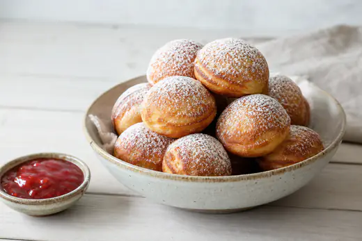 Gyldenbrune æbleskiver drysset med flormelis i en skål med syltetøj ved siden af