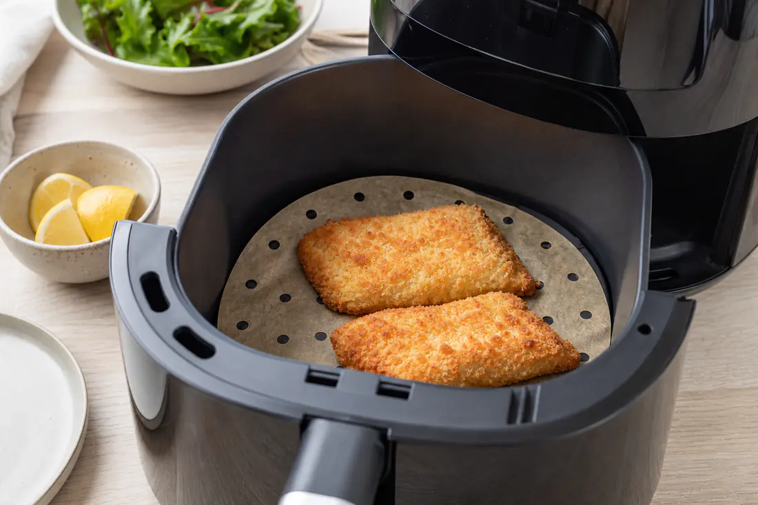 Airfryerkurv med et stykke perforeret bagepapir under to stykker paneret fisk