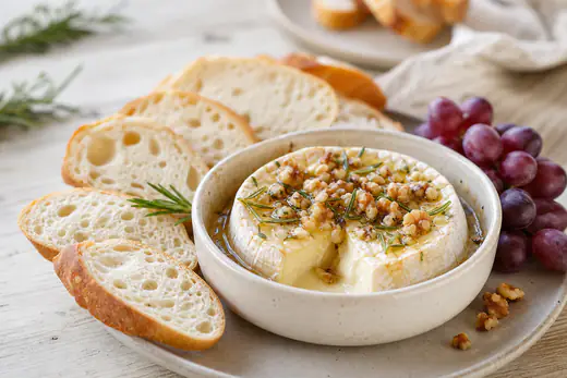 Bagt camembert med flydende midte, honning, rosmarin og valnødder i en lille form med baguette og druer ved siden af