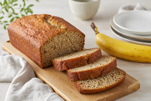 Banankage med gyldenbrun skorpe skåret i skiver på et bræt med en moden banan ved siden af