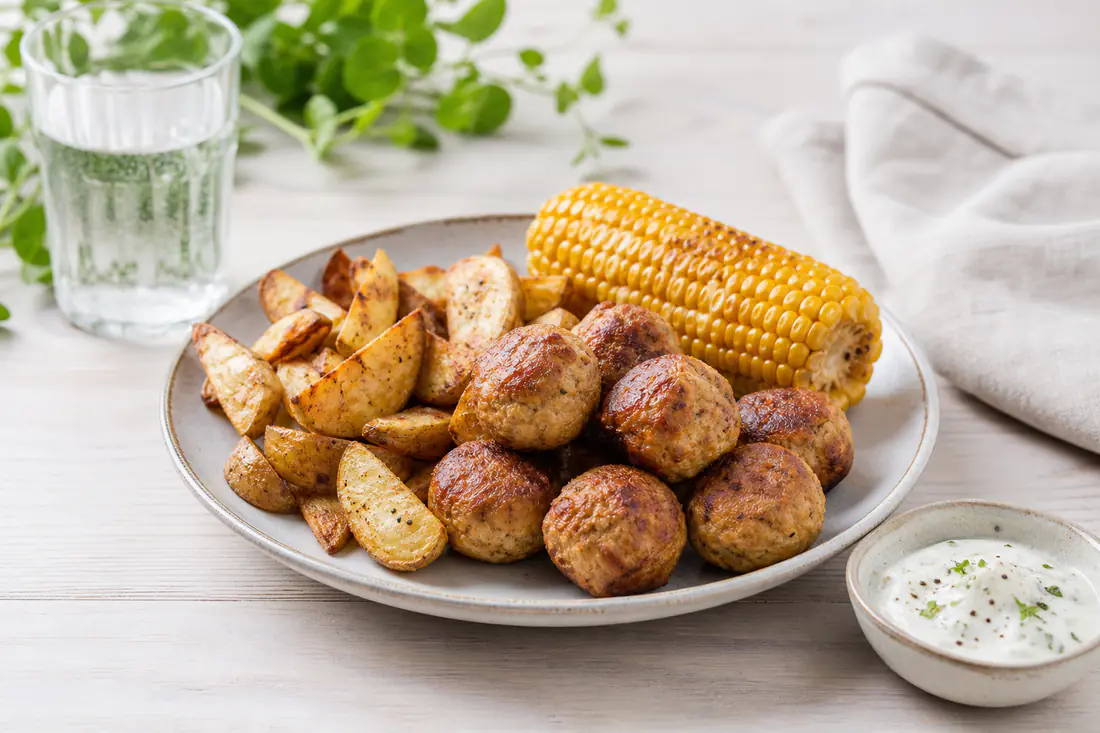 Tallerken med frikadeller, kartoffelbåde, majs og en lille skål dip på et lyst bord