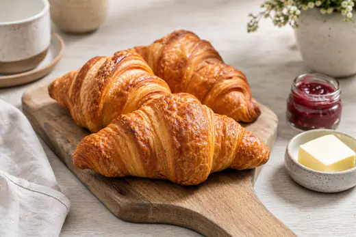 Gyldne, flaget croissanter på et træbræt med smør og marmelade ved siden af