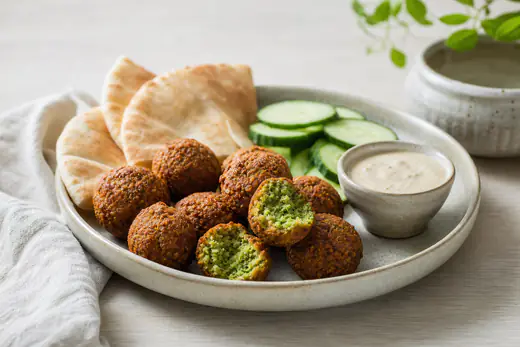 Sprøde falafler på en tallerken, hvor en er brækket over og viser grønt fyld, med tahinsauce, pitabrød og agurk