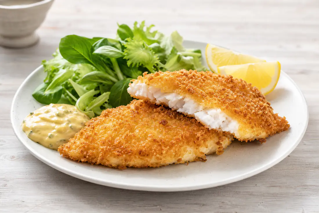 Sprøde, pankopanerede fiskefileter på en tallerken med citronbåde, remoulade og grøn salat