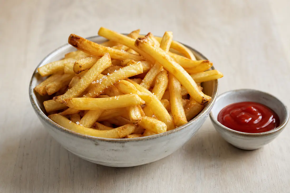 Gyldne, sprøde pommes frites drysset med salt i en skål med en lille skål ketchup ved siden af