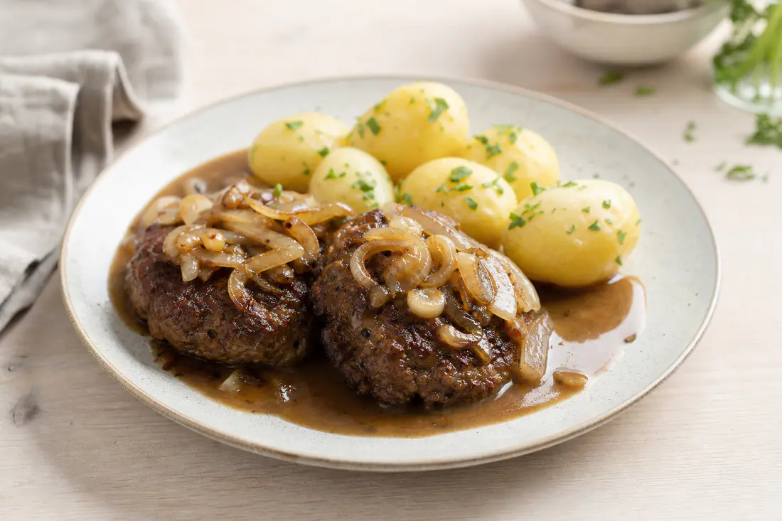 To brunstegte hakkebøffer med bløde løg, kogte kartofler og brun sovs på en tallerken