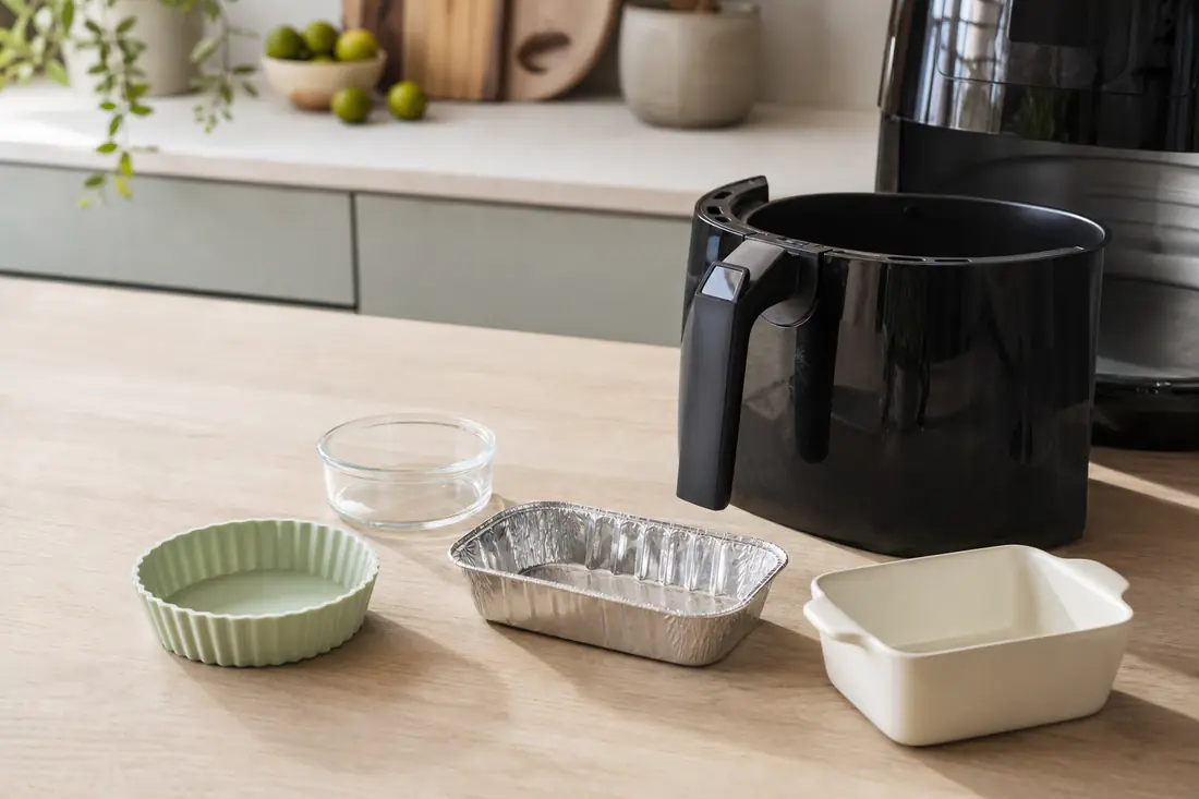Små forme i silikone, glas, aluminium og keramik på et bord ved siden af en åben airfryerkurv
