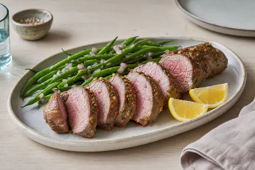 Skiveskåret lammefilet med rosa midte og gylden skorpe på en tallerken med grønne bønner og citronbåde