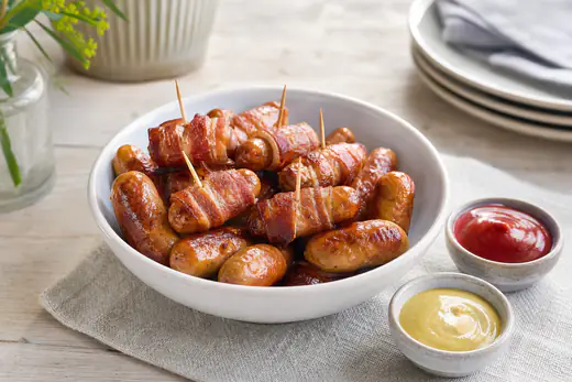 Små gyldenbrune cocktailpølser, nogle svøbt i bacon, i en skål med ketchup og sennep ved siden af