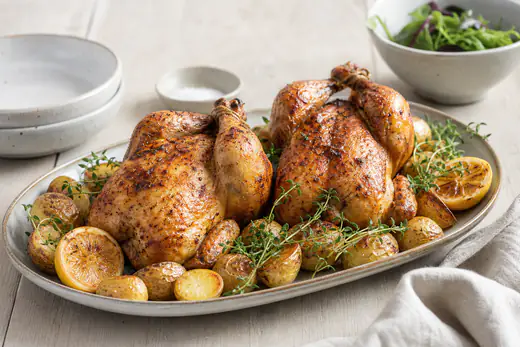 To hele gyldne poussiner med sprødt skind på et fad med citron, timian og kartofler