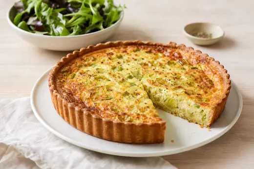 Gylden quiche med porre og ost i en lille tærteform, hvor et stykke er skåret ud og viser blød, sat æggemasse