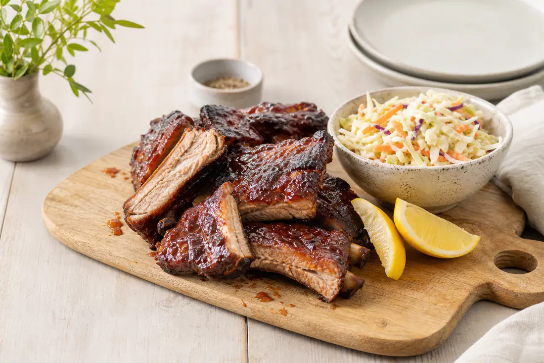 Glaserede spareribs skåret i stykker på et skærebræt med coleslaw og citronbåde ved siden af
