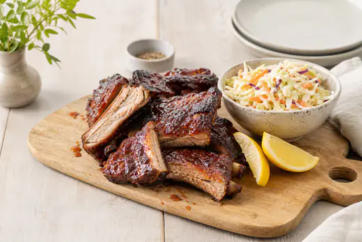 Glaserede spareribs skåret i stykker på et skærebræt med coleslaw og citronbåde ved siden af